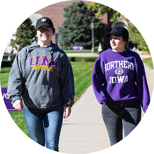 Two female UNI students wearing purple and gold walking across central campus